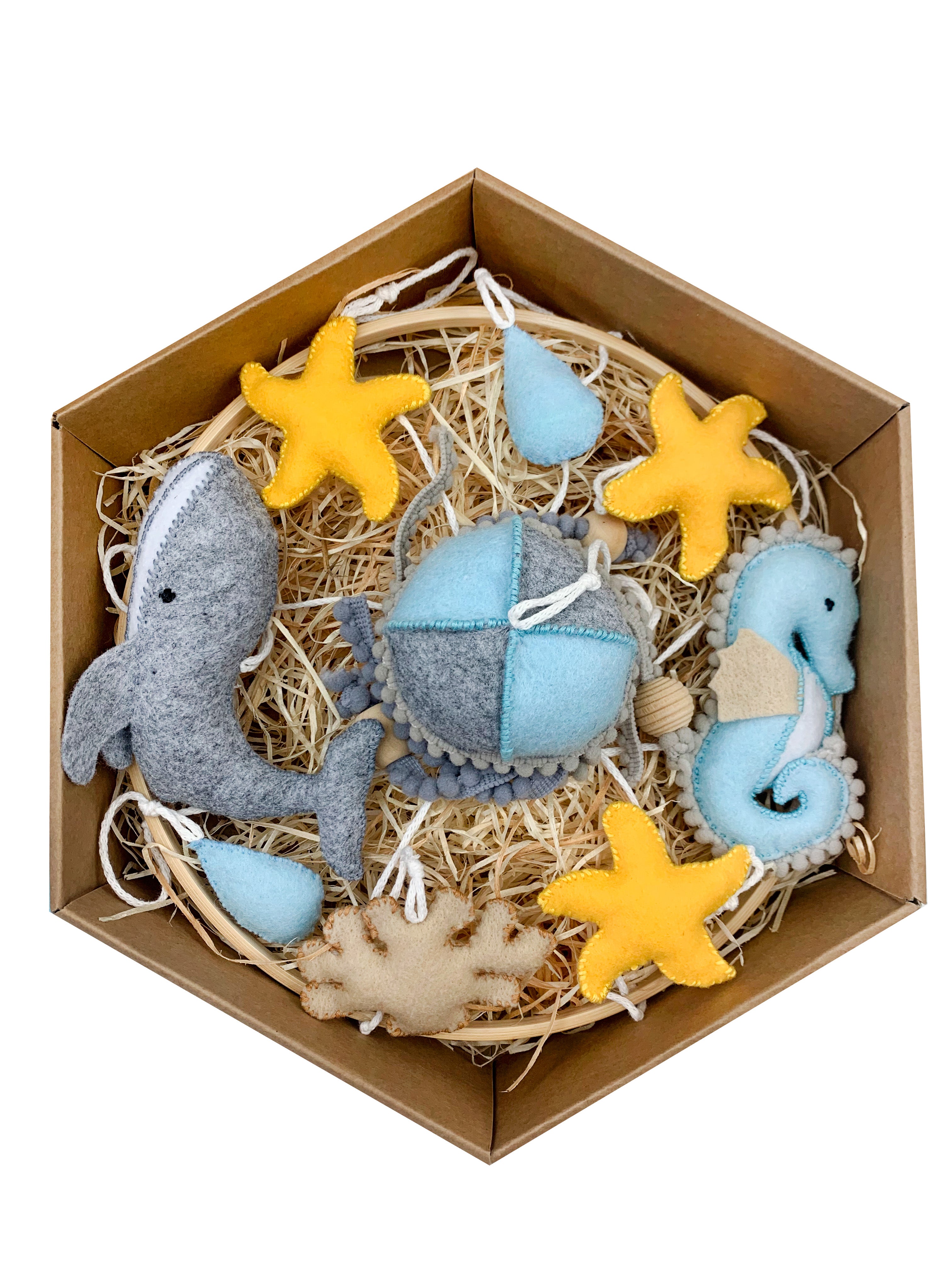 Dárkové balení dětského závěsného kolotoče s mořským motivem. V krabičce na podestýlce z dřevité vlny jsou ručně šité filcové dekorace: velryba, mořský koník, modré kapky, žluté hvězdice, korál a měkký textilní míček v modro-šedých tónech.