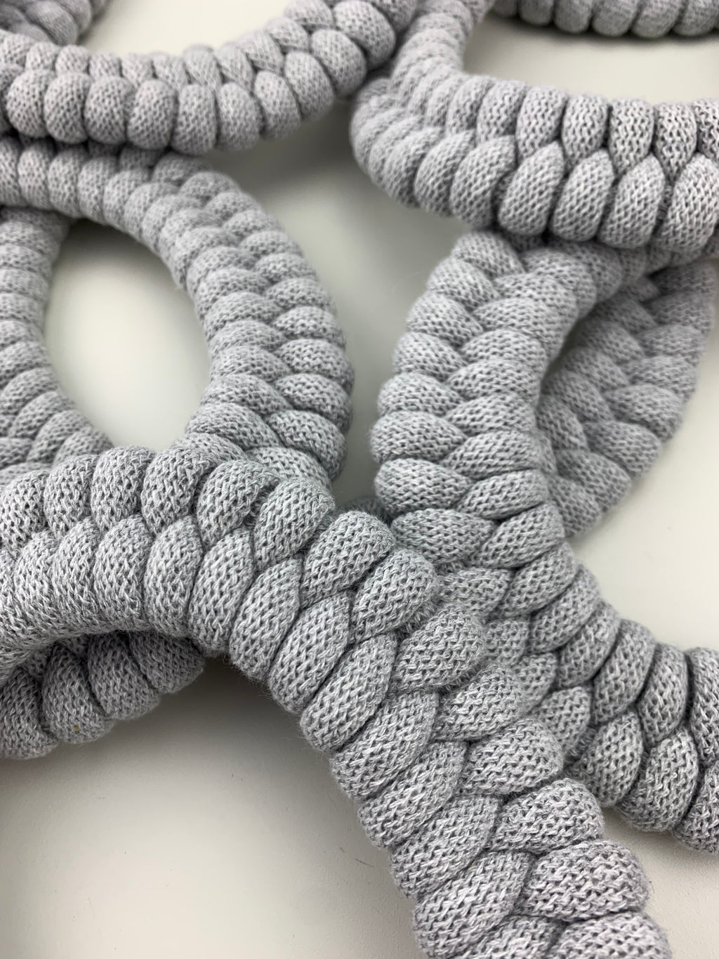Detailní pohled na několik světle šedých textilních náhrdelníků s hustým pravidelným pleteným vzorem. Náhrdelníky se překrývají a vytvářejí měkkou, strukturovanou kompozici na světlém podkladu.