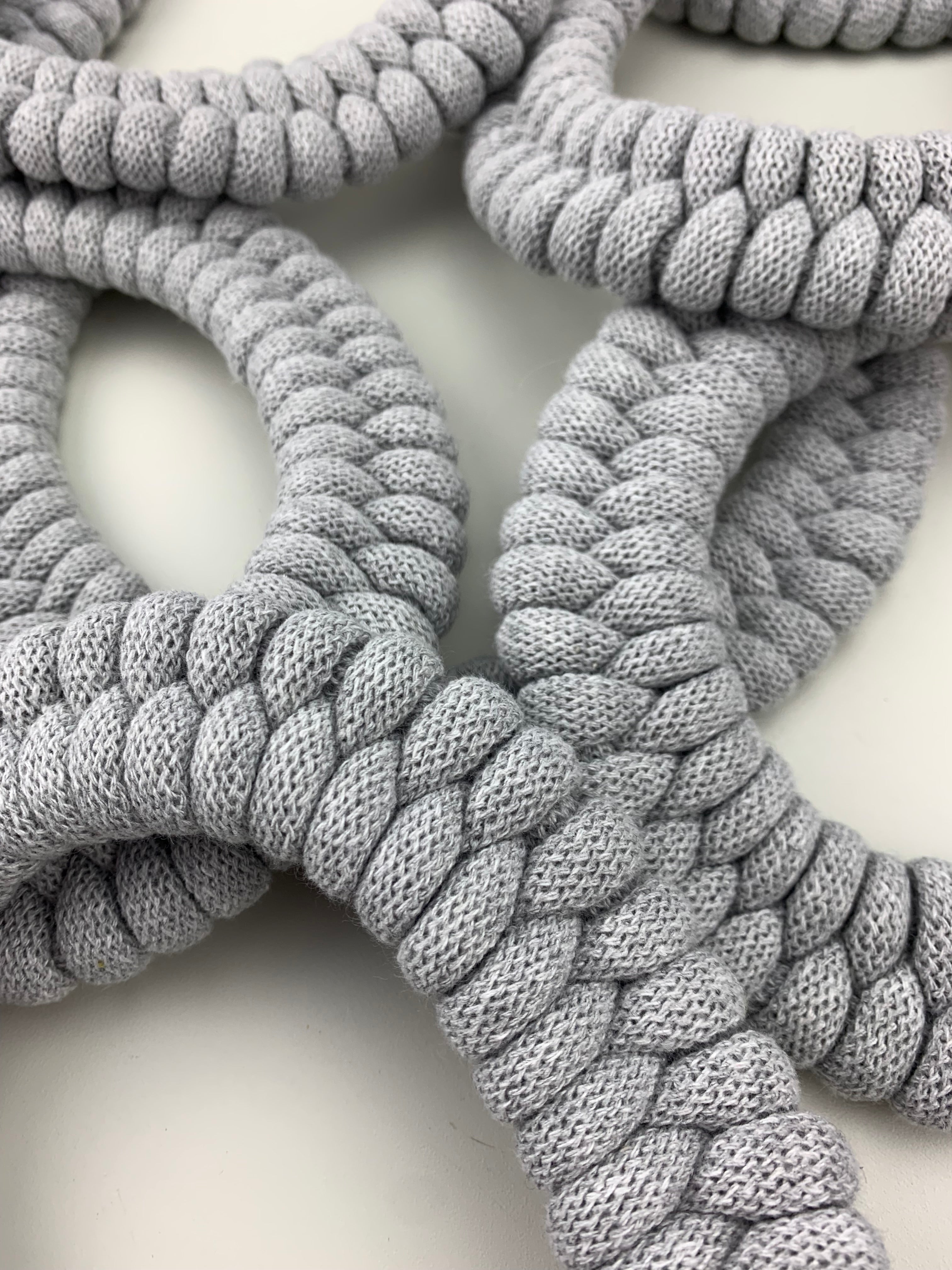Detailní pohled na několik světle šedých textilních náhrdelníků s hustým pravidelným pleteným vzorem. Náhrdelníky se překrývají a vytvářejí měkkou, strukturovanou kompozici na světlém podkladu.