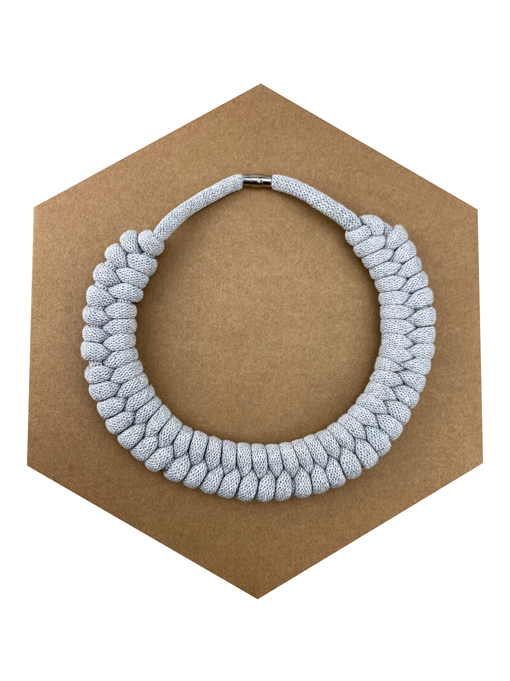 Ručně pletený náhrdelník ve světle šedé barvě, tvořený hustým pravidelným pleteným vzorem z měkké textilní šňůry. Náhrdelník je položený na hnědém šestiúhelníkovém podkladu a má kovové magnetické zapínání.