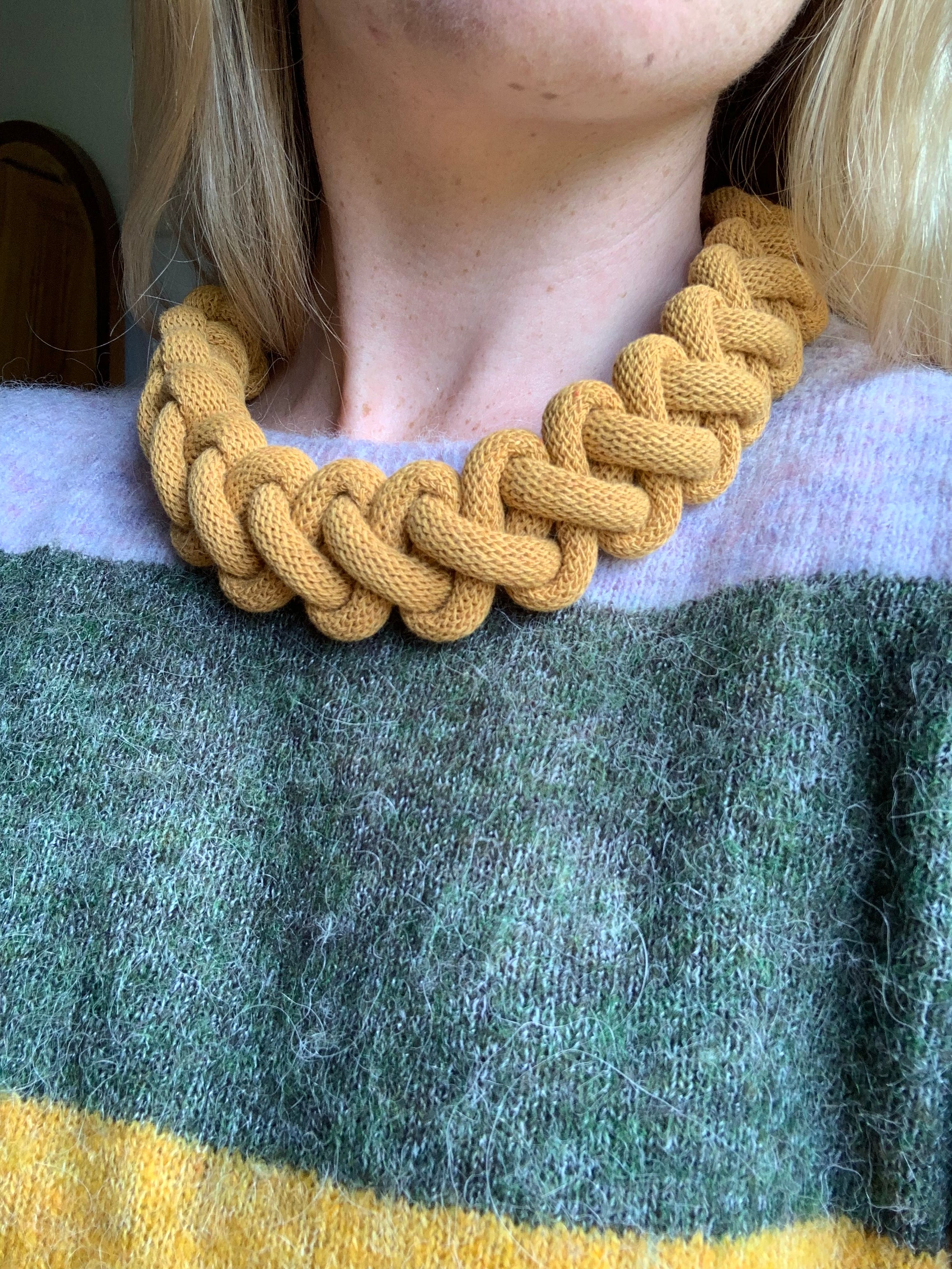 Detail ženy nesoucí kolem krku ručně pletený hořčicově žlutý náhrdelník z tlusté textilní šňůry. Náhrdelník má výrazný splétaný vzor a kontrastuje s měkkým svetrem v zelených a žlutých tónech.