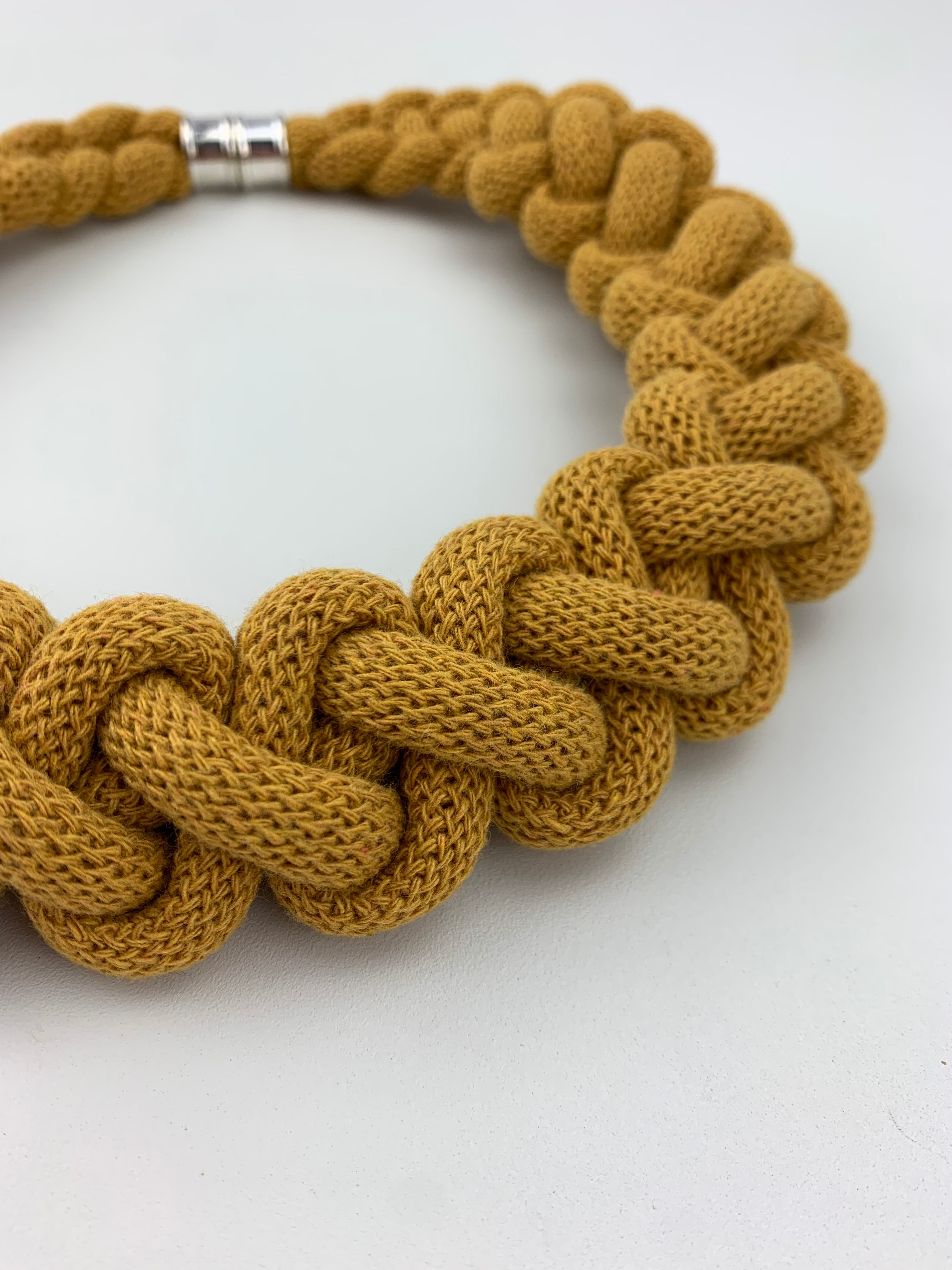 Detailní záběr na ručně pletený hořčicově žlutý náhrdelník z měkké textilní šňůry, tvořený výrazným spleteným vzorem. V pozadí je vidět kovové magnetické zapínání, náhrdelník leží na čistě bílém povrchu.