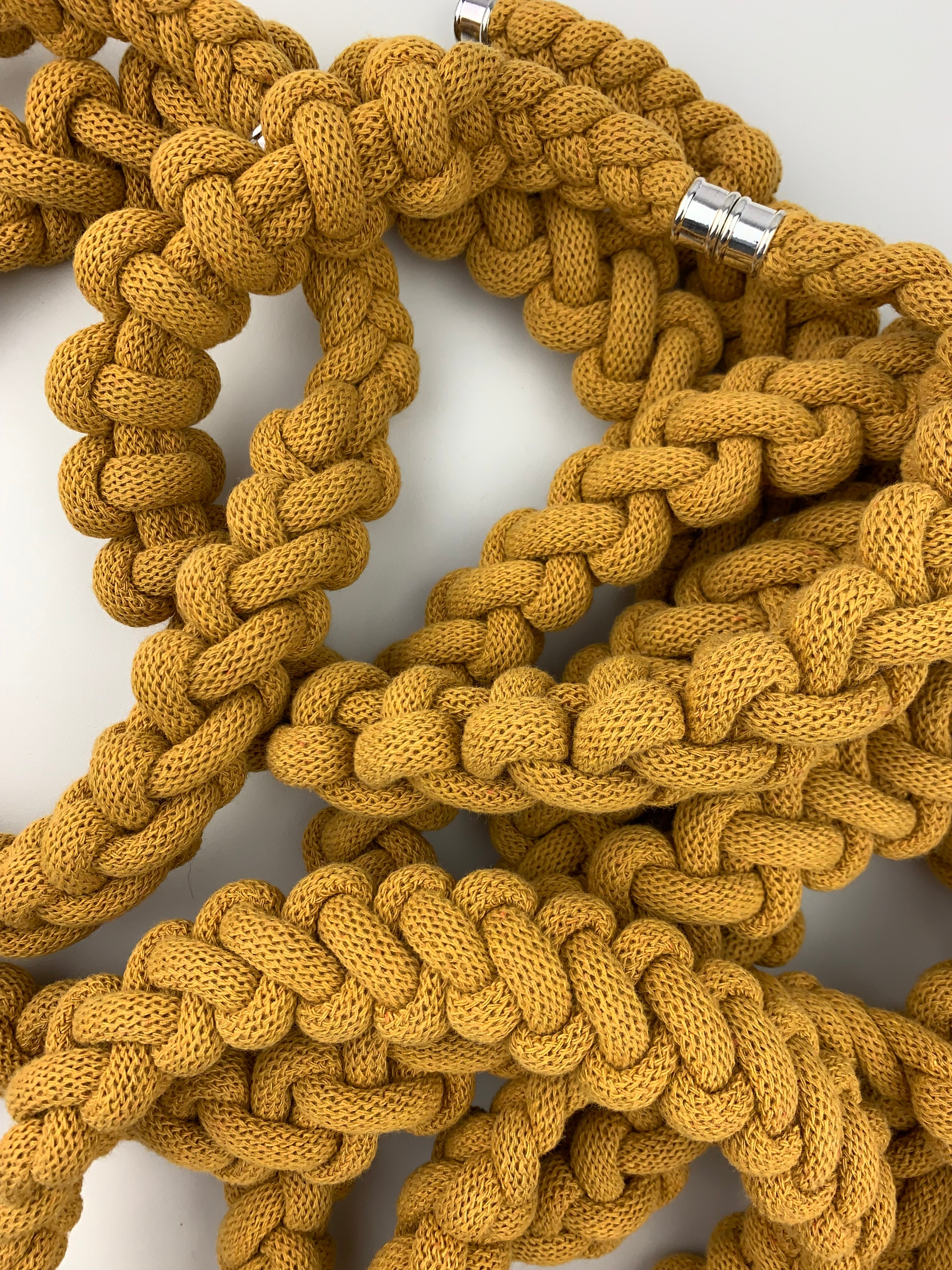 Skupina několika ručně pletených hořčicově žlutých náhrdelníků z měkké textilní šňůry, složených do sebe a překrývajících se. Jsou vytvořeny z robustního spleteného vzoru a doplněny kovovým magnetickým zapínáním. Náhrdelníky leží na světlém podkladu.