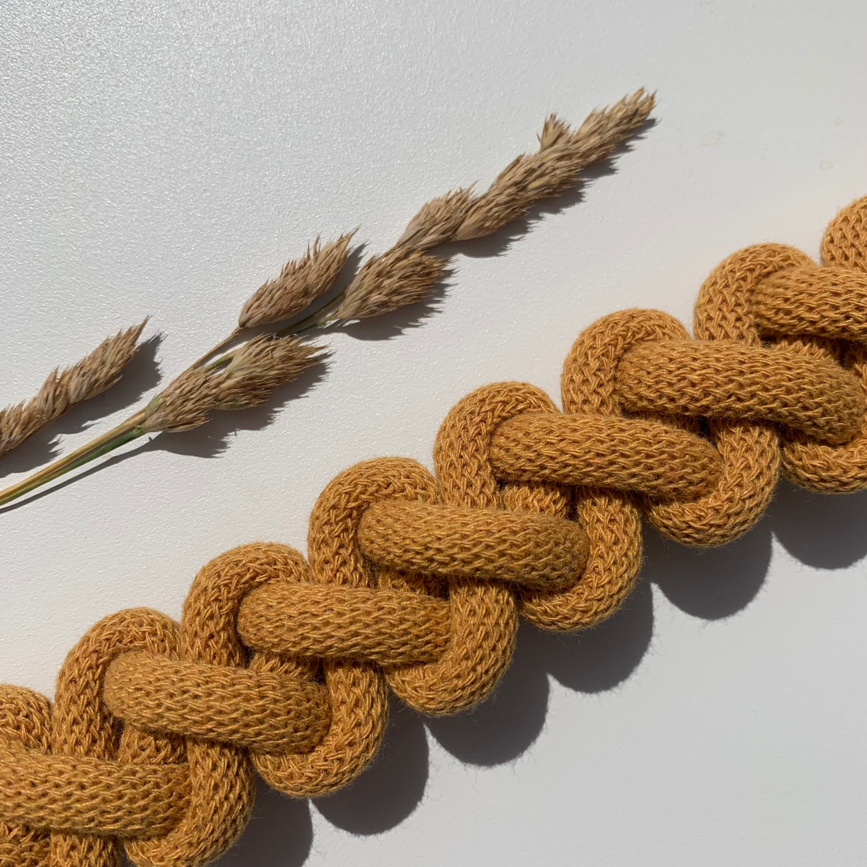 Detail spleteného hořčicově žlutého textilního náhrdelníku položeného na bílém povrchu. Vedle něj leží dvě sušené traviny, které vytvářejí přírodní a minimalistickou kompozici.