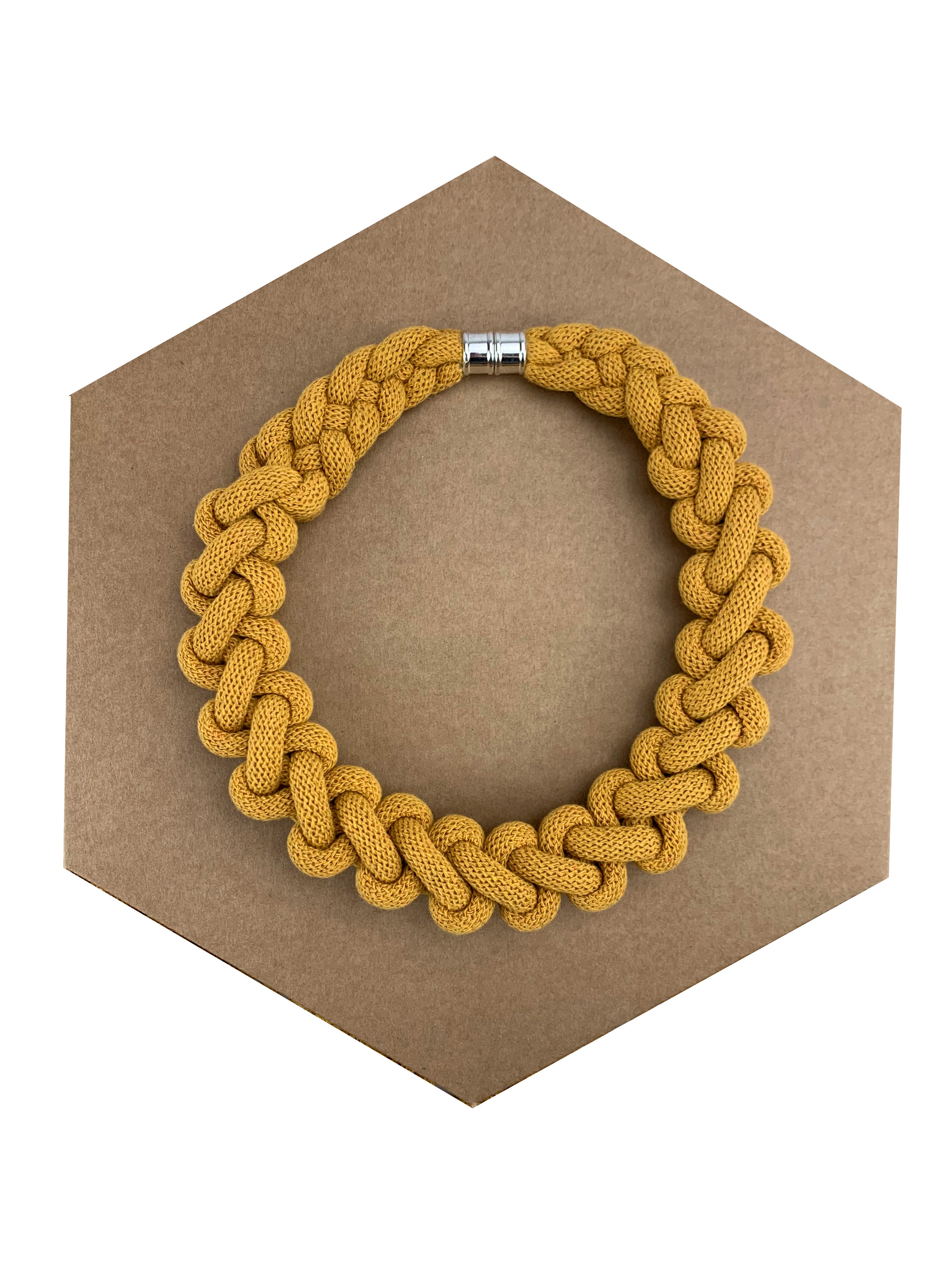 Ručně pletený náhrdelník v teplé hořčicově žluté barvě, vytvořený z měkké textilní šňůry do výrazného spleteného vzoru. Náhrdelník je položený na hnědém šestiúhelníkovém podkladu a má kovové magnetické zapínání.
