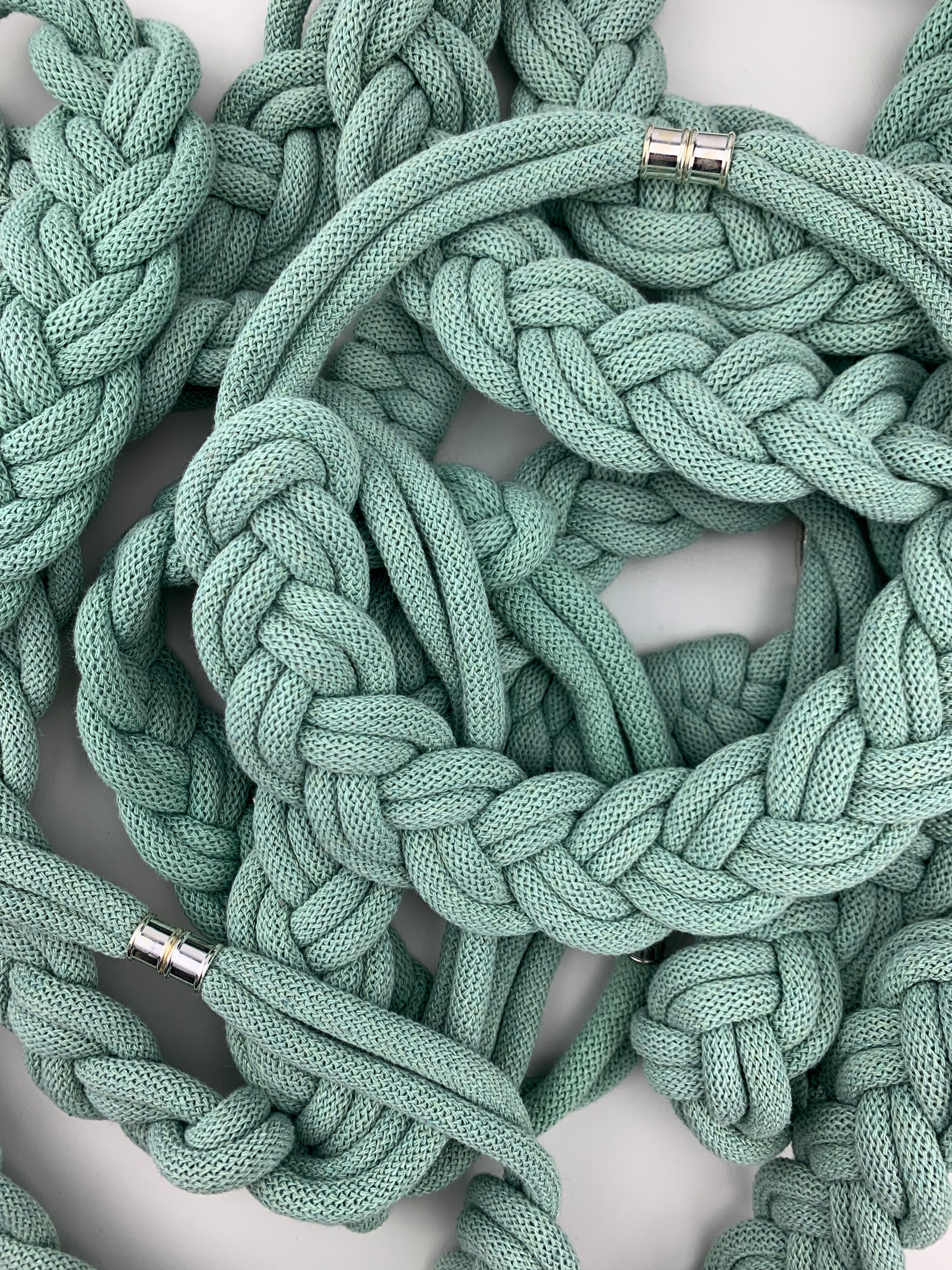 Spletená hromada několika mentolově zelených textilních náhrdelníků s robustním pleteným vzorem. Náhrdelníky se překrývají a vytvářejí hustou strukturu, na několika místech jsou viditelná kovová magnetická zapínání.