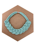Ručně pletený náhrdelník v jemné mentolově zelené barvě, vytvořený z měkké textilní šňůry spletené do výrazného uzlového vzoru. Náhrdelník je položený na hnědém šestiúhelníkovém podkladu a má kovové magnetické zapínání.