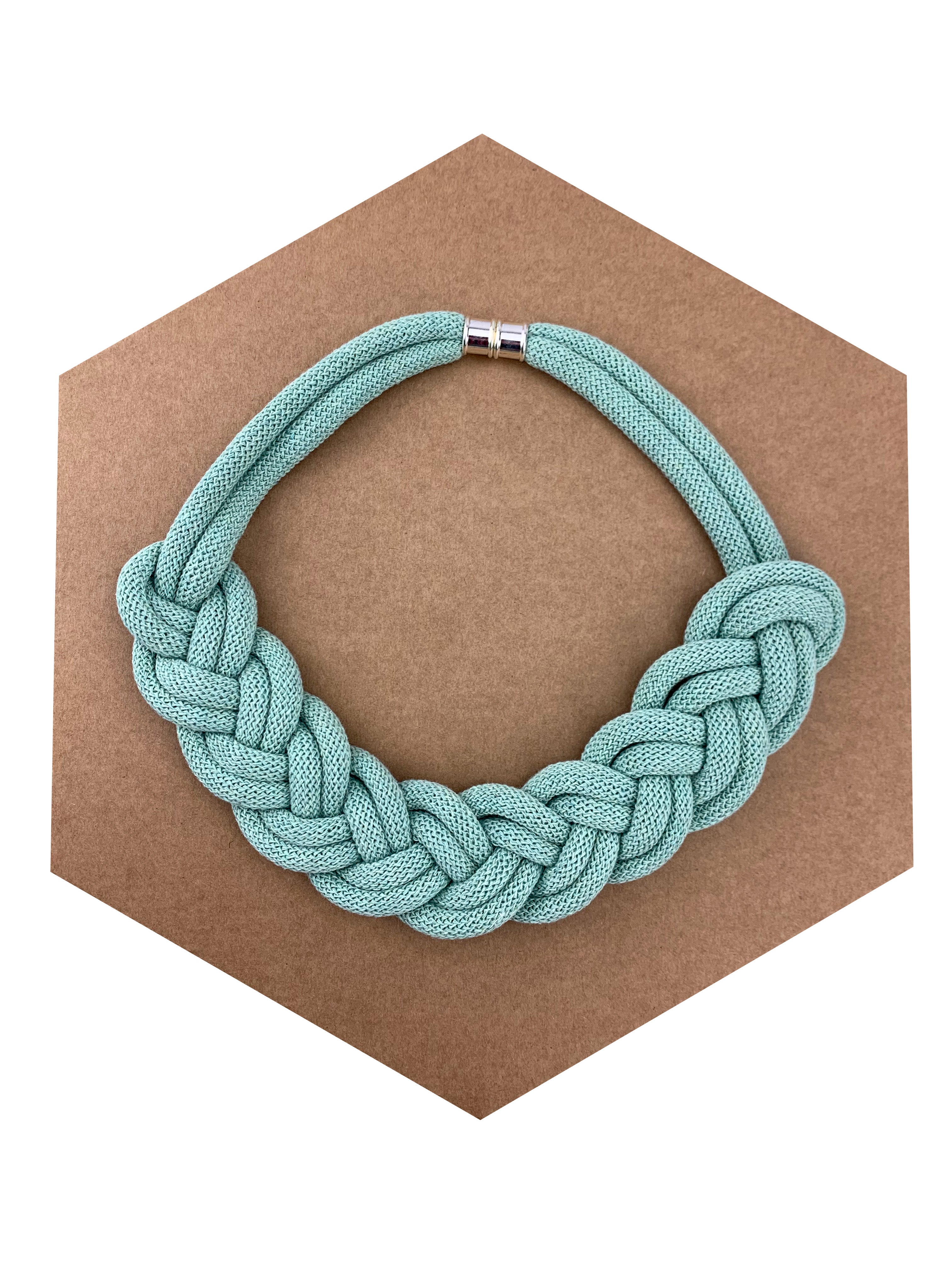 Ručně pletený náhrdelník v jemné mentolově zelené barvě, vytvořený z měkké textilní šňůry spletené do výrazného uzlového vzoru. Náhrdelník je položený na hnědém šestiúhelníkovém podkladu a má kovové magnetické zapínání.