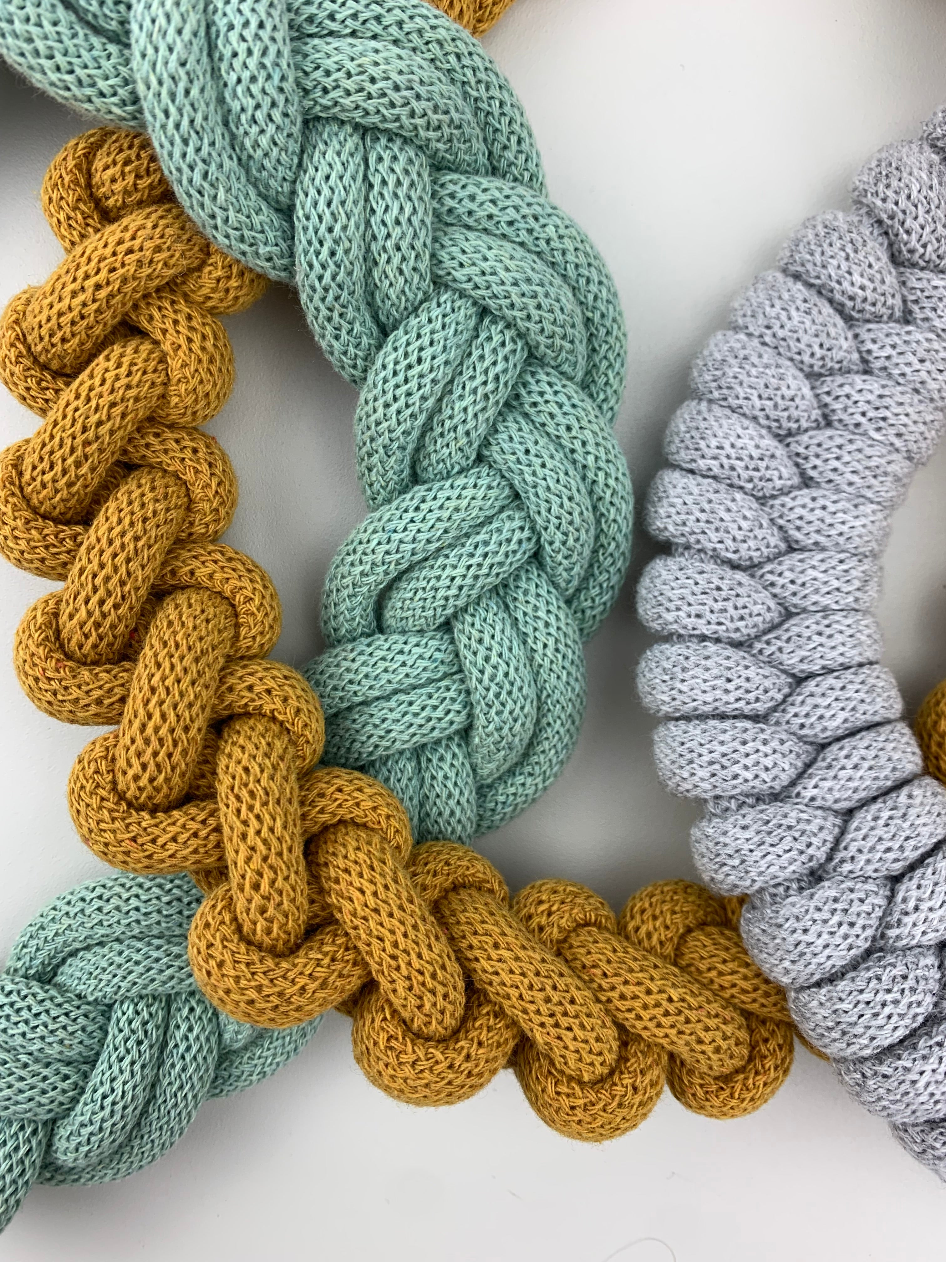 Detail tří ručně pletených textilních náhrdelníků v různých barvách – hořčicově žluté, mentolově zelené a světle šedé. Náhrdelníky mají výrazný spletený vzor a leží vedle sebe na světlém podkladu.